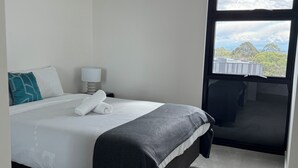 Ein ordentlich bezogenes Bett mit weißen Leinen und einem aufgerollten Handtuch, eine Nachttischlampe und ein Fenster mit Blick auf Bäume und Gebäude.