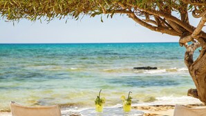 A beach scene with two wooden chairs and a small round table under a tree, overlooking the sea. - Deluxe Two-bedroom Apartment (Trachoni)