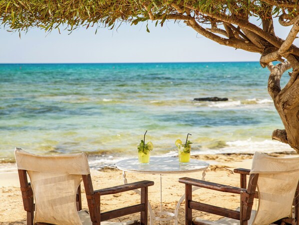 Escena de playa con dos sillas de madera y una mesa redonda pequeña bajo un árbol, con vista al mar.