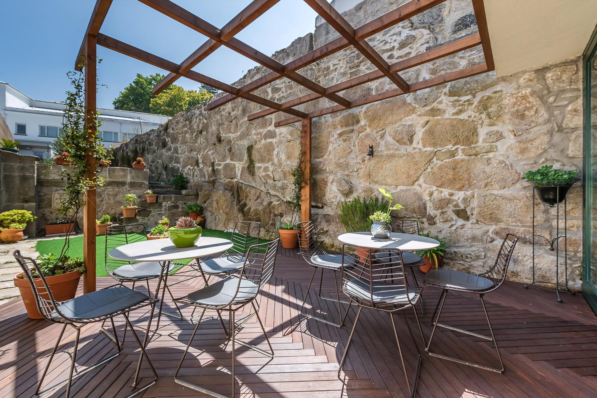 A covered patio with wooden flooring, metal chairs, and tables, surrounded by stone walls and potted plants.