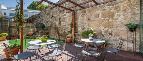 A covered patio with wooden flooring, metal chairs, and tables, surrounded by stone walls and potted plants.