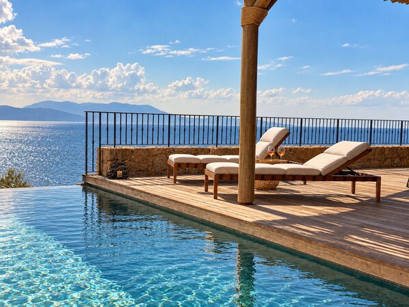 A wooden deck with lounge chairs overlooking a clear blue sea and mountains.