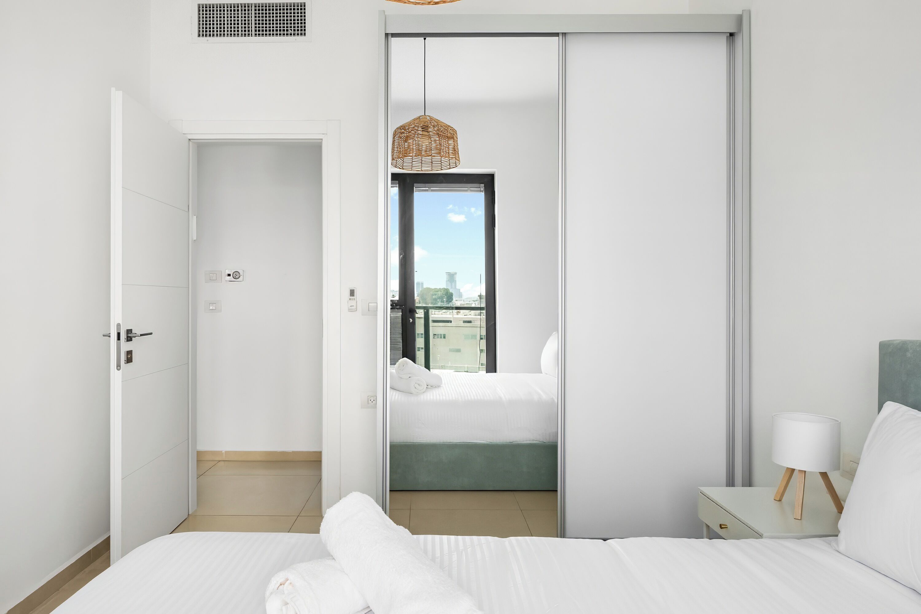 A modern hotel room with a bed, a nightstand, a lamp, and a view of the city through a sliding door.