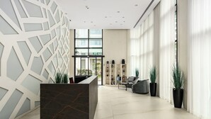 En moderne lobby med en marmorreception, et geometrisk vægdesign og et siddeområde med sofa og potteplanter.