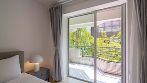 Un dormitorio con cama, una mesita de noche con lámpara y vistas a un balcón con vegetación.