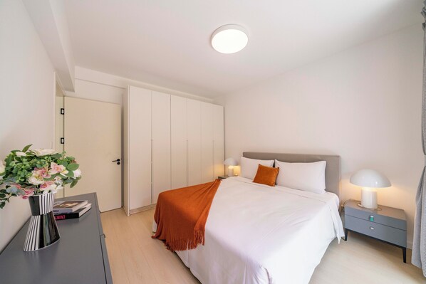 A modern bedroom with a bed, bedside tables, a nightstand, a door, and a vase with flowers. - Tyms Bright 2BR 1BA Apt Xuhui Center (Shanghai)