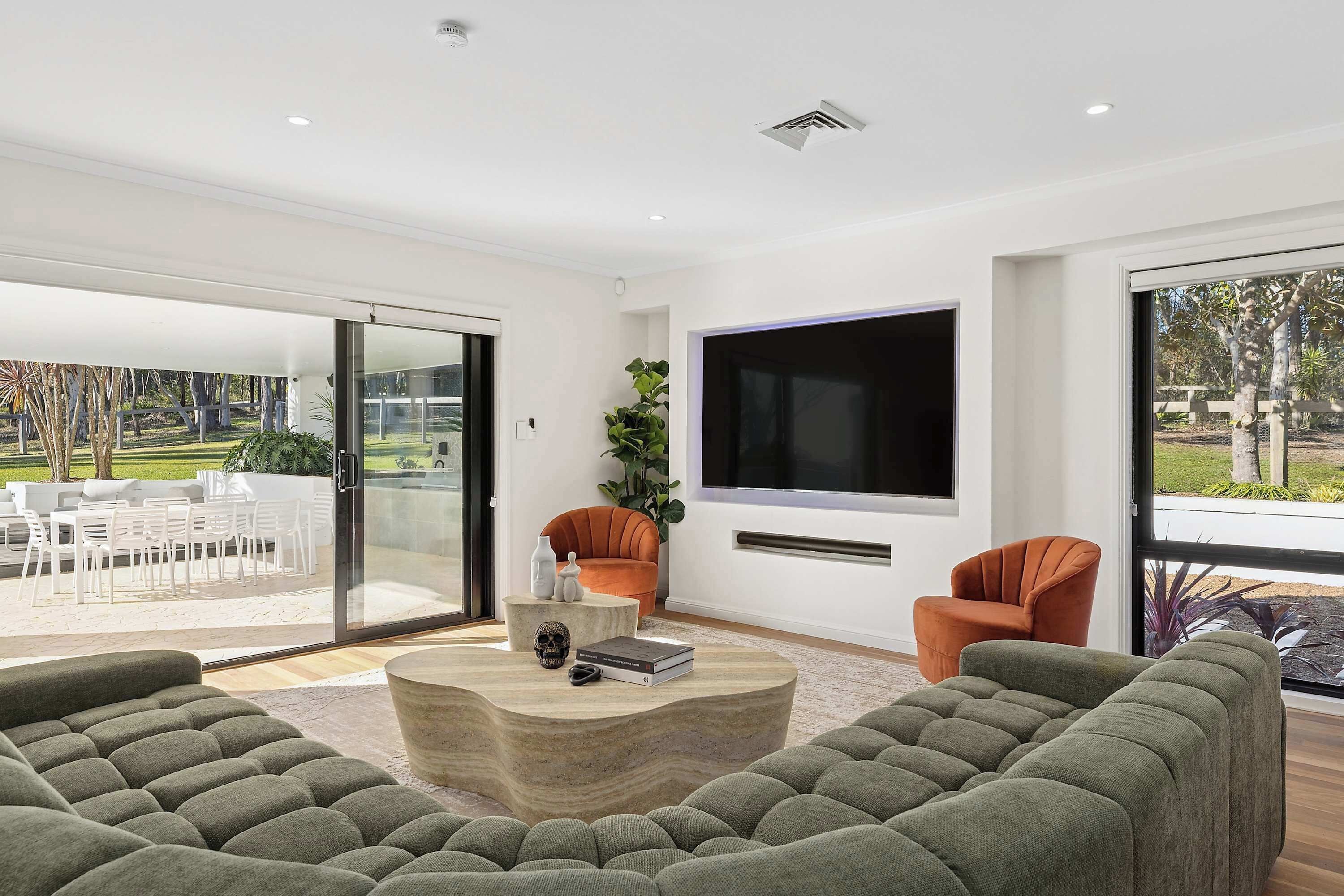 A modern living room with a large TV, green sofas, and a glass door leading to an outdoor patio.