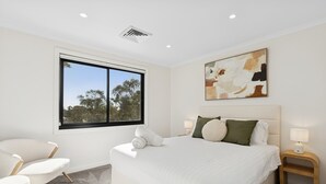 Un dormitorio moderno con una cama grande, una ventana con vistas a árboles, un cuadro en la pared y una mesita de noche con una lámpara.