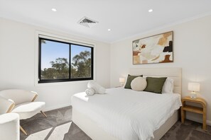 A modern bedroom with a large bed, a window with a view of trees, a painting on the wall, and a small bedside table with a lamp.