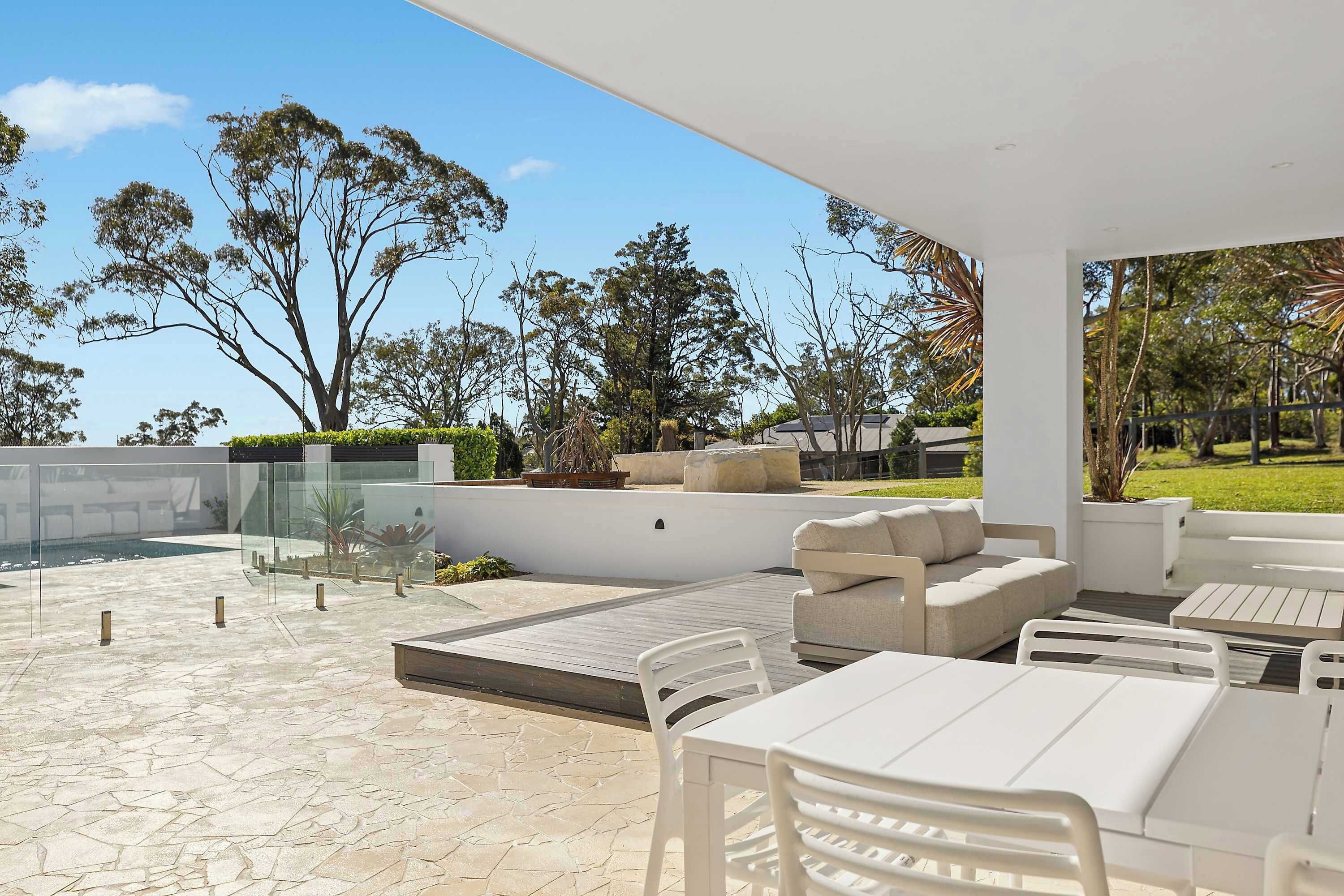 Une terrasse extérieure avec un sol en pierre, des meubles blancs et une vue sur des arbres et un bâtiment en arrière-plan.