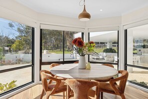 A modern dining area with a round table and six chairs, large windows offering a view of the outdoors, and a hanging pendant light.