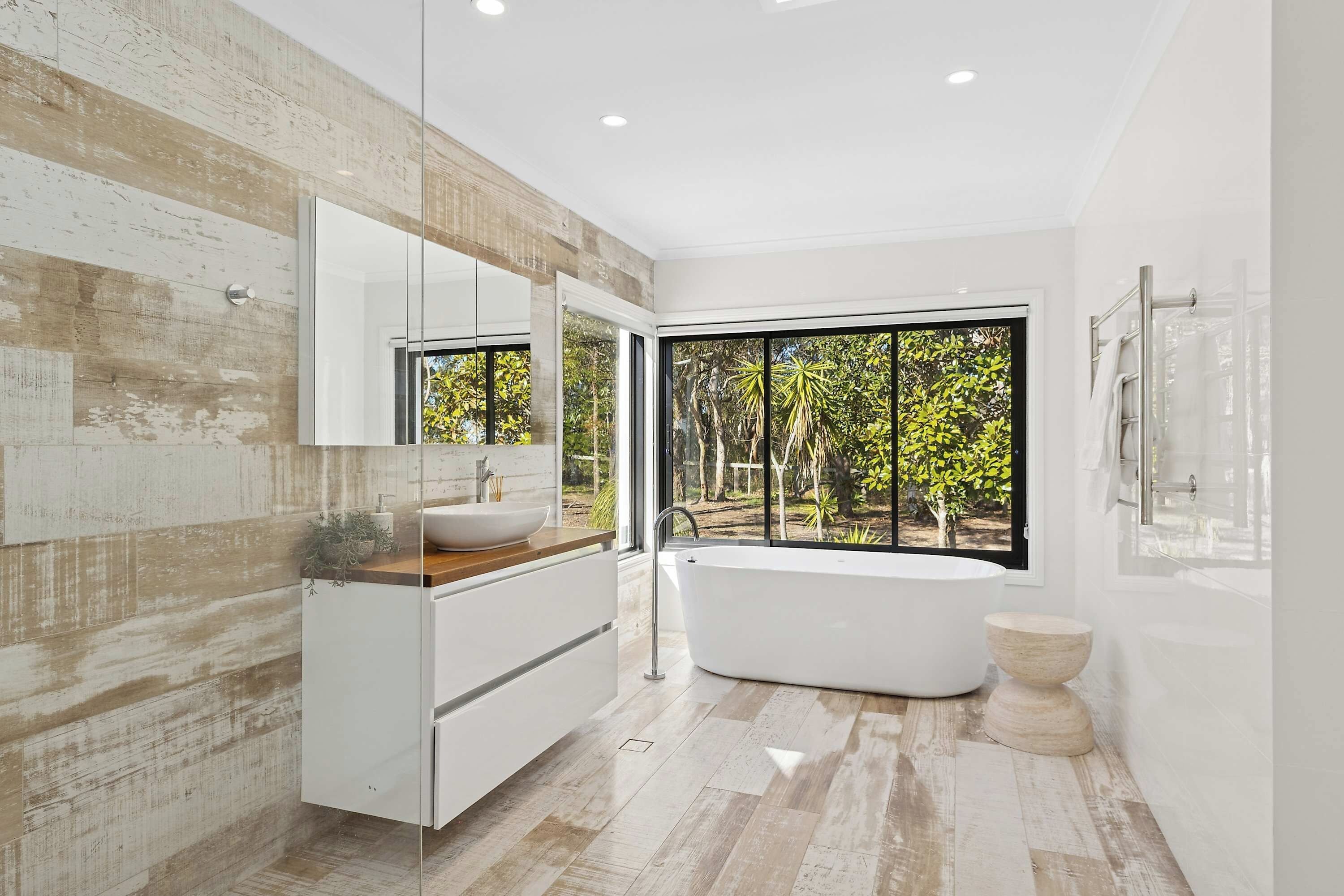 A modern bathroom with a freestanding bathtub, a large mirror, and a view of trees through a large window.