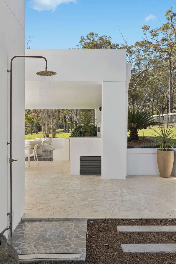 A modern outdoor patio with a stone floor, a white wall, a large potted plant, and a view of a garden with trees and a lawn.