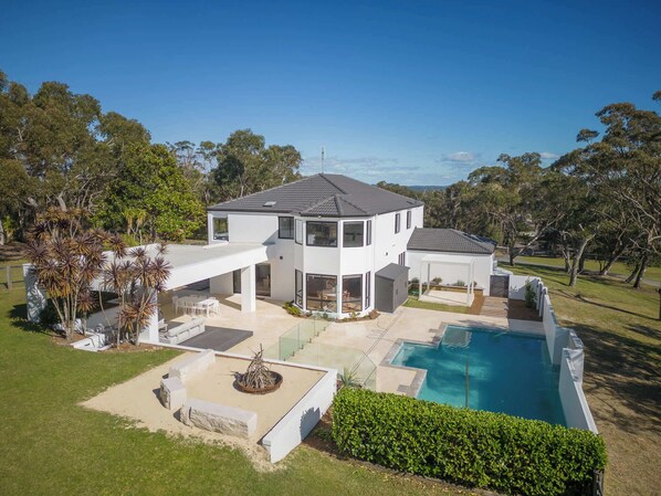 A modern two-story house with a large swimming pool, surrounded by a well-maintained lawn and trees.