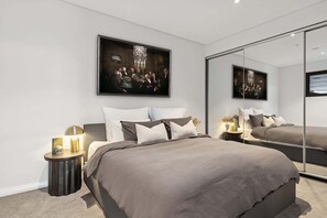 A modern bedroom with a large bed, a bedside table with a lamp, and a large mirror reflecting the room. - Artful Lakefront Apartment | Avoca, Sleeps 4, EVC (Avoca Beach)