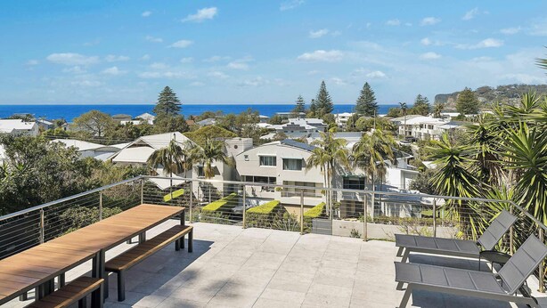 A terrace with a wooden table and benches, overlooking a coastal town with houses and trees.