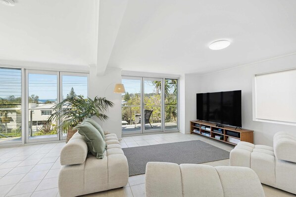 A modern living room with a large TV, comfortable seating, and a view through large windows.