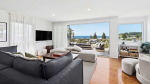 A modern living room with a grey sofa, white armchairs, and a flat-screen TV. A large window offers a view of the ocean and hills.