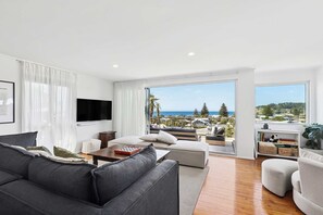 Una sala de estar moderna con un sofá gris, sillones blancos y un televisor de pantalla plana. Un ventanal ofrece vistas al océano y a las colinas.