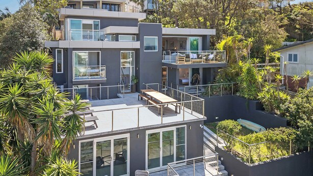 A modern multi-story house with large windows, outdoor seating, and a staircase. - Ocean Views Every Room. 100m to Beach. Sleeps 13 (North Avoca)