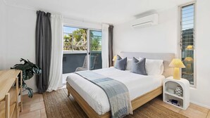 A bedroom with a bed, bedside lamp, and a small white nightstand. - Ocean Views Every Room. 100m to Beach. Sleeps 13 (North Avoca)