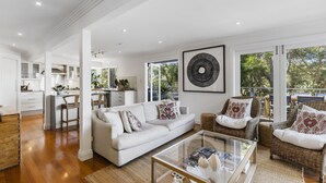 A spacious living area with a white sofa, wicker chairs, and a glass coffee table. A kitchen is visible in the background with white cabinets and a dining area.