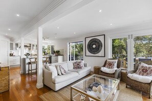 Un amplio salón con un sofá blanco, sillas de mimbre y una mesa de centro de cristal. Se observa una cocina al fondo con armarios blancos y una zona de comedor.