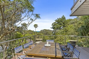 A wooden deck with striped chairs and a table, surrounded by lush greenery and a clear blue sky.