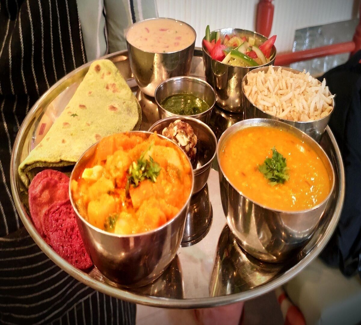 Comida tradicional india servida en una bandeja de acero inoxidable con diferentes platos, incluyendo un tazón de dal naranja, un trozo de roti, un tazón de verduras mixtas y un recipiente pequeño de chutney.