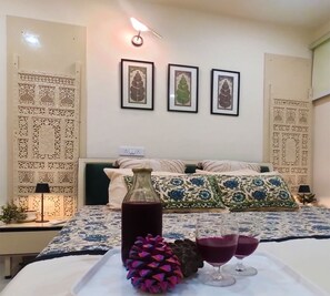 A bedroom with a bed, decorative pillows, a bedside table with a lamp, a wall with three framed pictures, and a tray with a bottle and glasses. - Aahaar (Dharamshala)