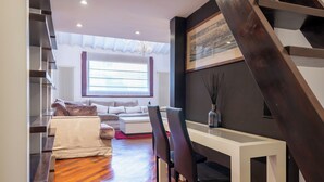 Een moderne woonkamer met een trap, een bank, een eettafel met stoelen en een groot raam met jaloezieën.