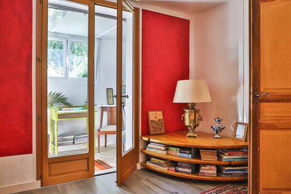 Una habitación con una pared roja, una puerta de madera, una estantería, una lámpara y una pequeña mesa con una silla.
