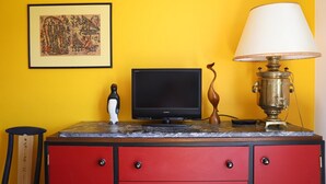 A room with a yellow wall, a framed artwork, a television, a lamp, and a red cabinet with drawers.