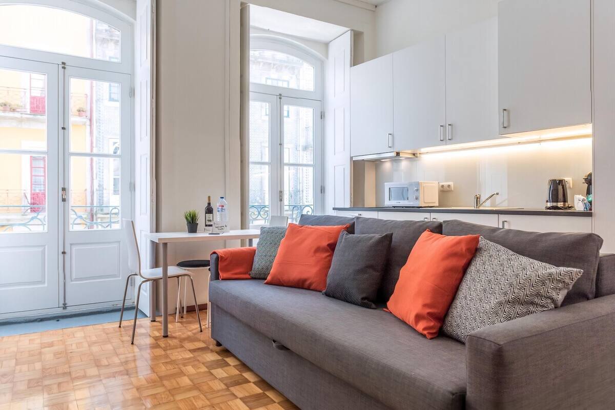 A modern living room with a grey sofa, a wooden floor, and a kitchen area with white cabinets and a microwave.