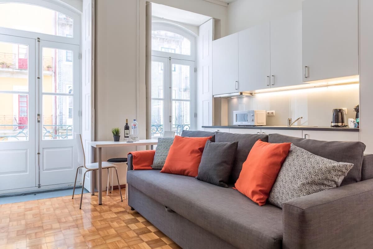 A modern living room with a grey sofa, a wooden floor, and a kitchen area with white cabinets and a microwave.