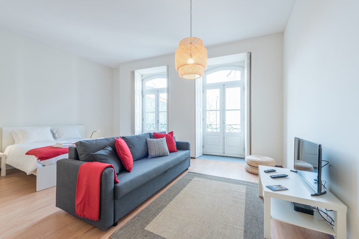 A modern living room with a grey sofa, red cushions, a woven light fixture, a flat-screen TV, and a large window.