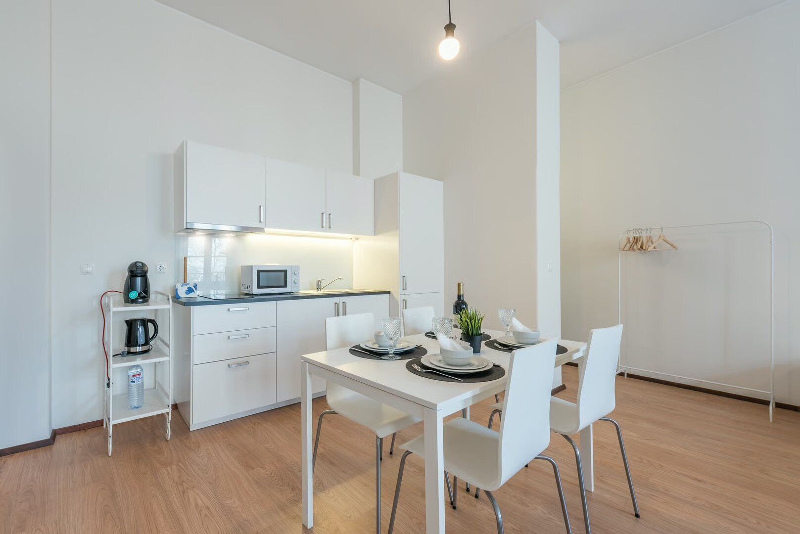 A modern kitchen with white cabinets, a microwave, and a kettle. There is a dining table set with plates, glasses, and a bottle.