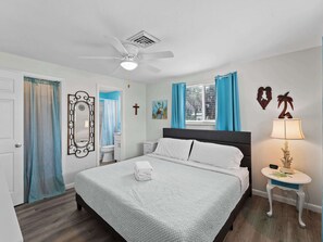 A bedroom with a bed, a ceiling fan, a window with curtains, a nightstand, a lamp, and a wall with a cross and decorative items.