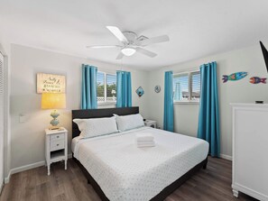 Une chambre à coucher avec un lit, des tables de chevet, une lampe, un ventilateur de plafond et une fenêtre avec des rideaux.