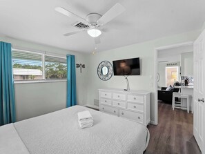 A bedroom with a bed, a ceiling fan, a TV, a dresser, and a window with curtains.