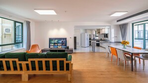 A modern living room with a wooden sofa, a dining table with chairs, a television, and a kitchen in the background.