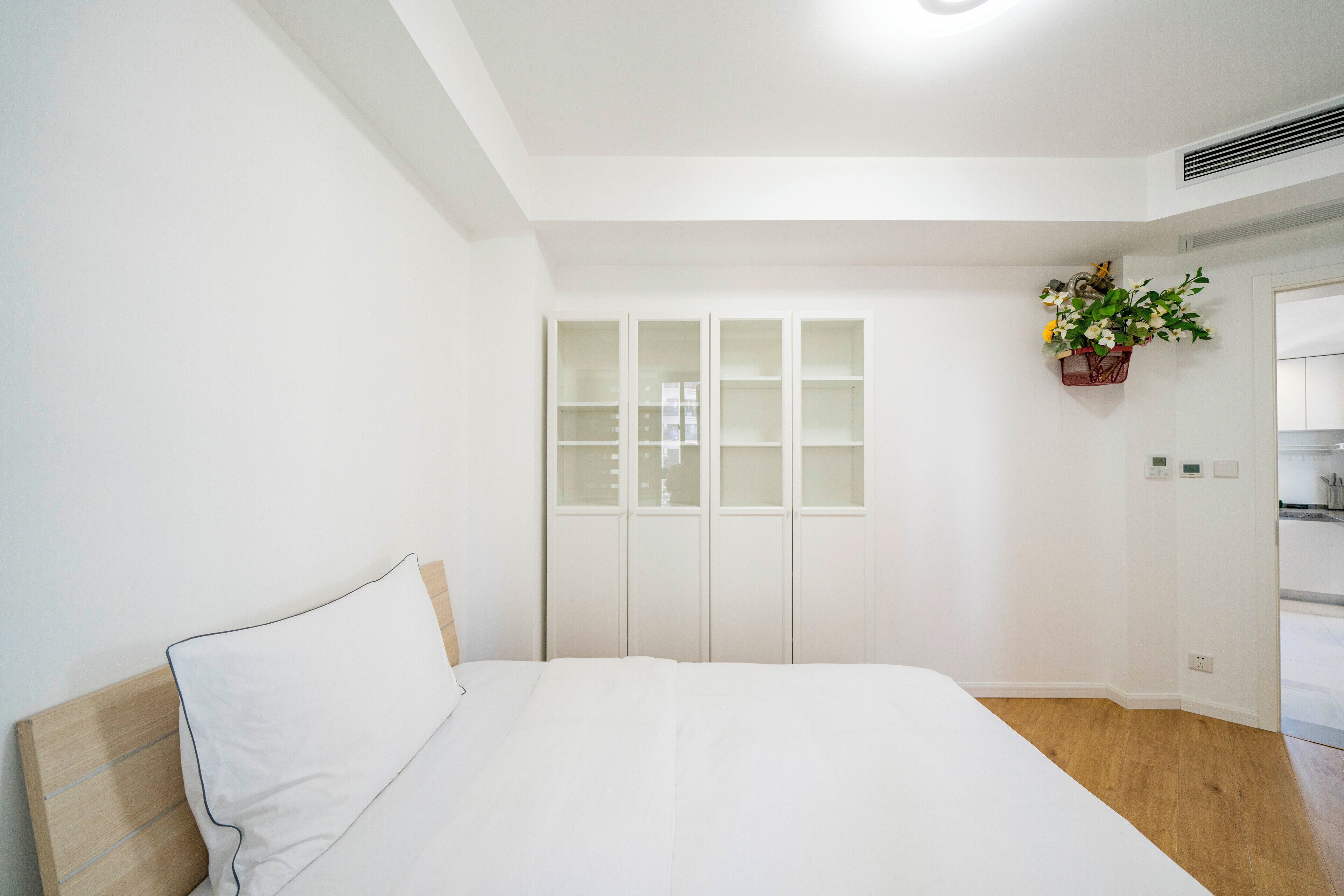 Een slaapkamer met een bed, een houten hoofdbord, een witte muur, een witte kledingkast en een kleine plant in pot.