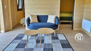 A cozy room with a blue sofa, two wooden tables, and a checkered rug. A clock is mounted on the wall, and a bed is visible in the adjacent room.