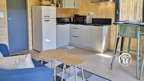 A compact kitchen with a refrigerator, microwave, and stove, a dining area with a table and chairs, and a sofa in a room with wooden walls and a door.