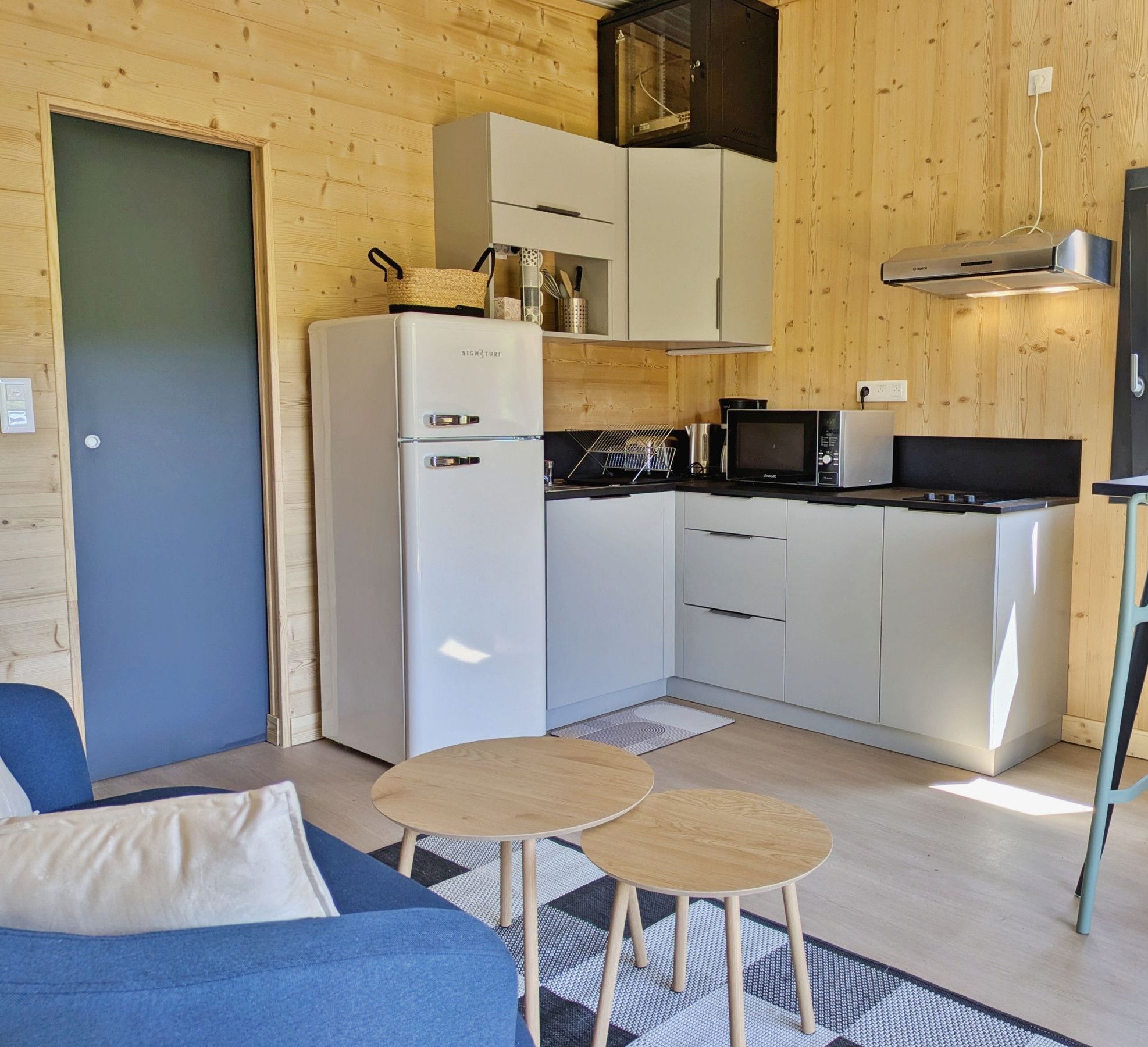 Cocina compacta con refrigerador, microondas y estufa, un comedor con mesa y sillas, y un sofá en una habitación con paredes de madera y una puerta.
