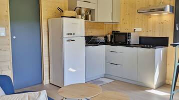 Cocina compacta con refrigerador, microondas y estufa, un comedor con mesa y sillas, y un sofá en una habitación con paredes de madera y una puerta.