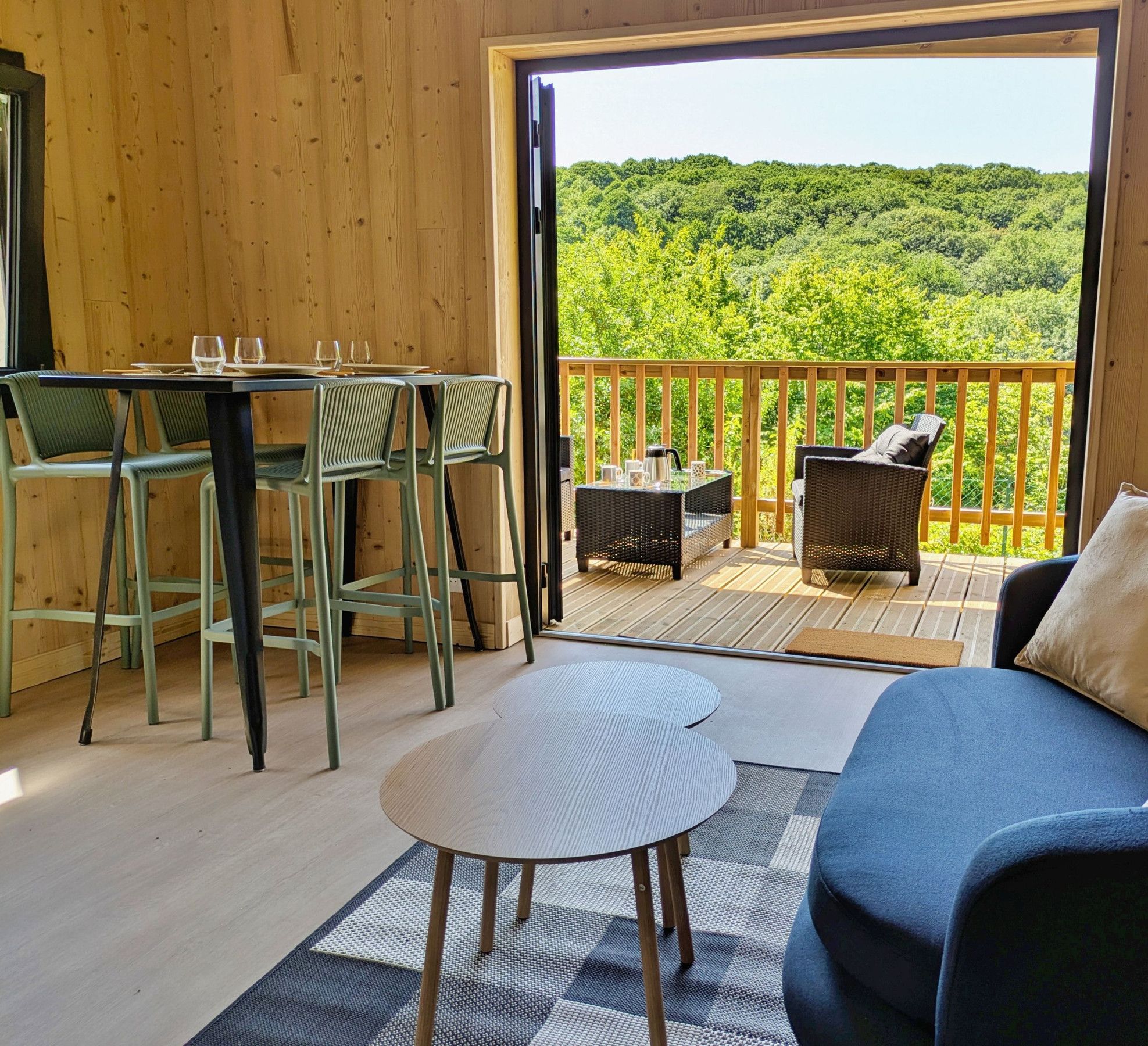 Une pièce chaleureuse au décor intérieur en bois, un balcon avec vue sur une forêt, un coin repas avec des chaises et une table, et un canapé agrémenté de coussins.