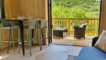 Une pièce chaleureuse au décor intérieur en bois, un balcon avec vue sur une forêt, un coin repas avec des chaises et une table, et un canapé agrémenté de coussins.