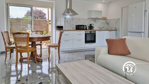 Eine moderne Küche mit weißen Schränken, einem eingebauten Backofen und einer Mikrowelle. Es gibt einen Essbereich mit einem runden Tisch und Stühlen, sowie ein weißes Sofa mit einem braunen Kissen.