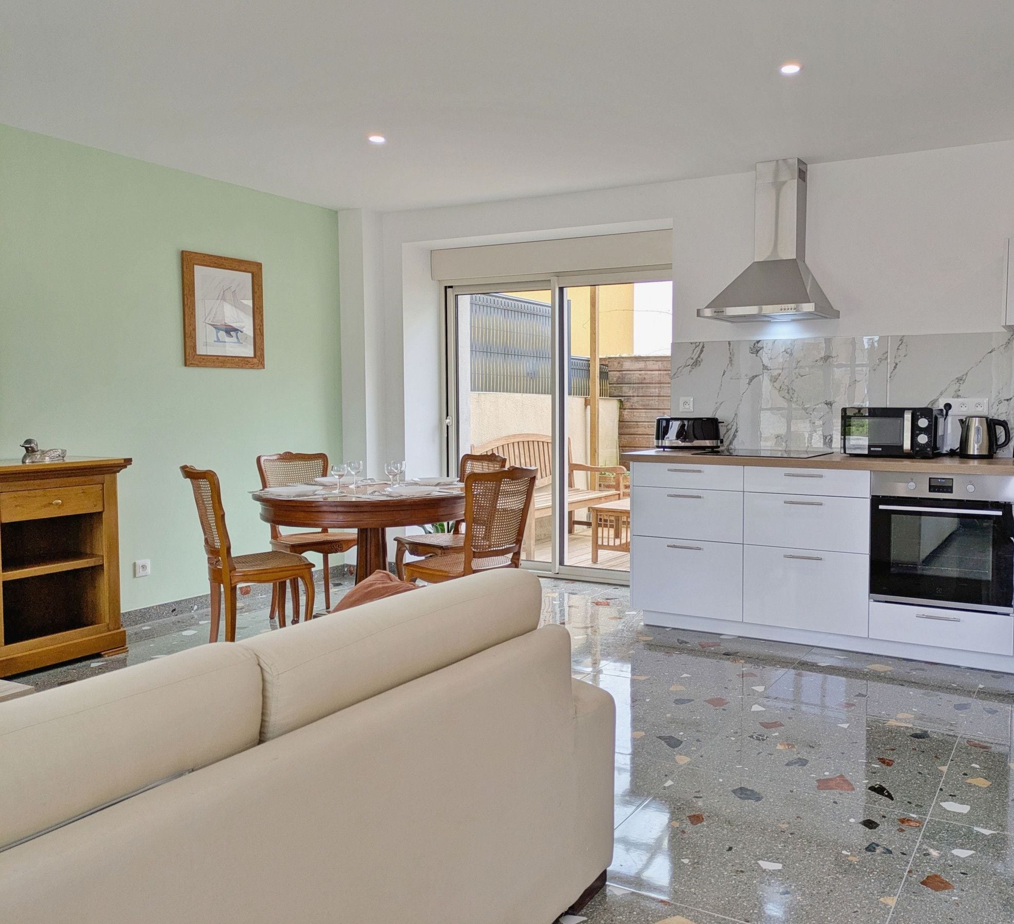Una cocina moderna con armarios blancos, horno empotrado y cafetera. Hay un comedor con una mesa y sillas de madera, y un sofá en la sala de estar.